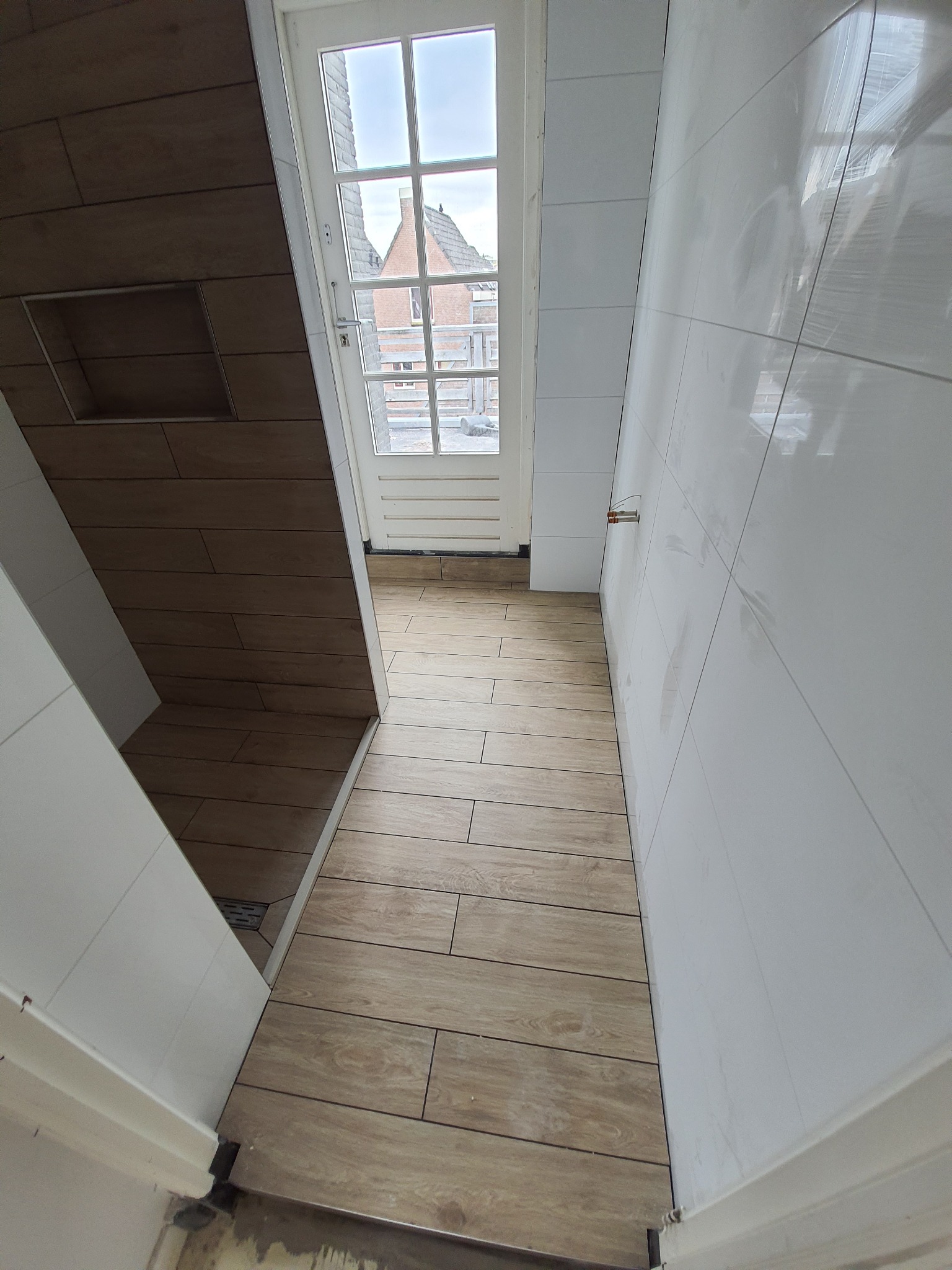 Wykończona łazienka z drewnopodobnymi płytkami na podłodze i ścianach, białe płytki na ścianie, widoczny odpływ liniowy i okno z widokiem na budynki.