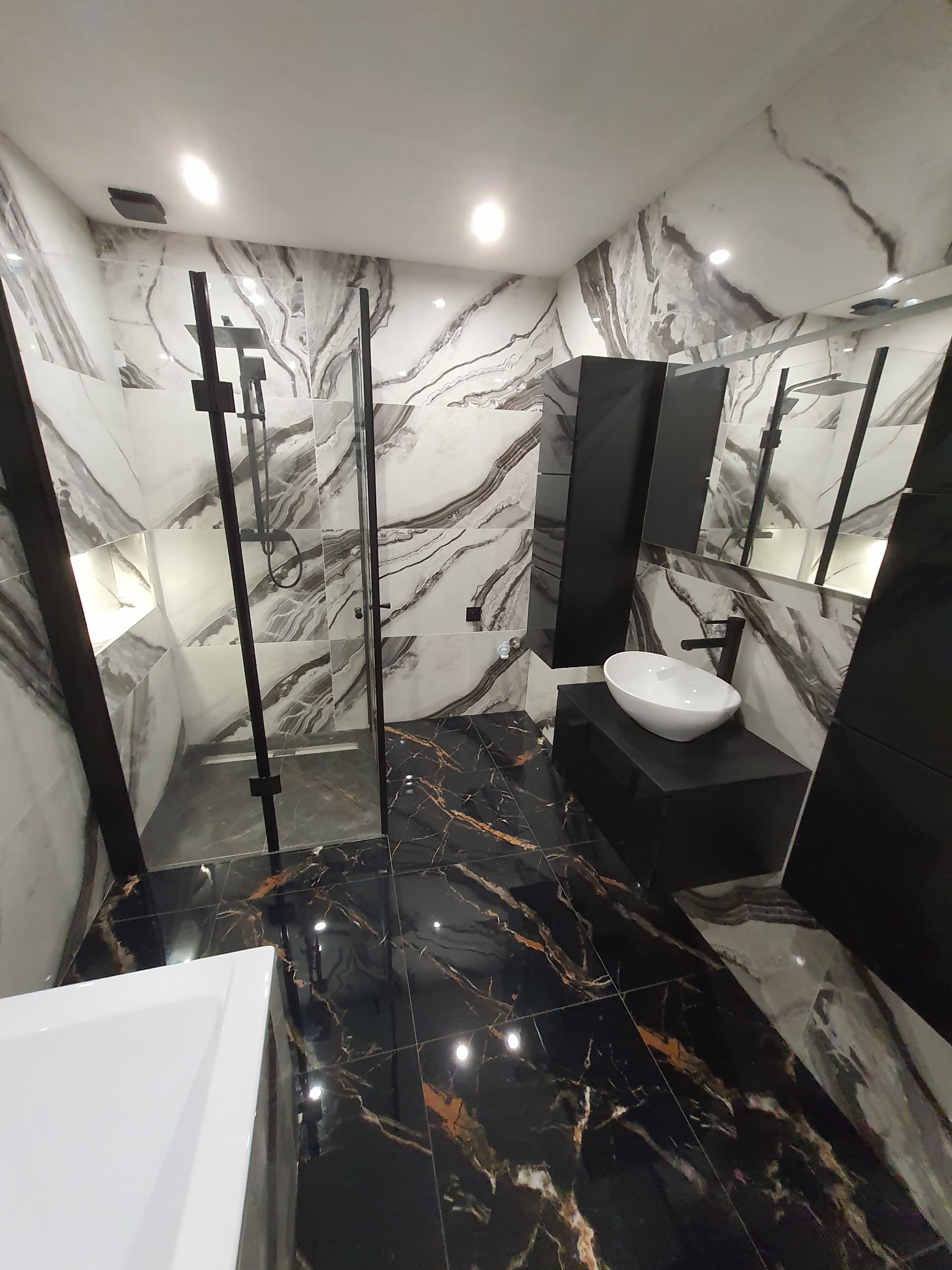 Wykończona łazienka w czarno-białej kolorystyce z marmurowymi płytkami na ścianach i podłodze, kabina prysznicowa typu walk-in z czarnymi ramami i armaturą, umywalka nablatowa na czarnej szafce.