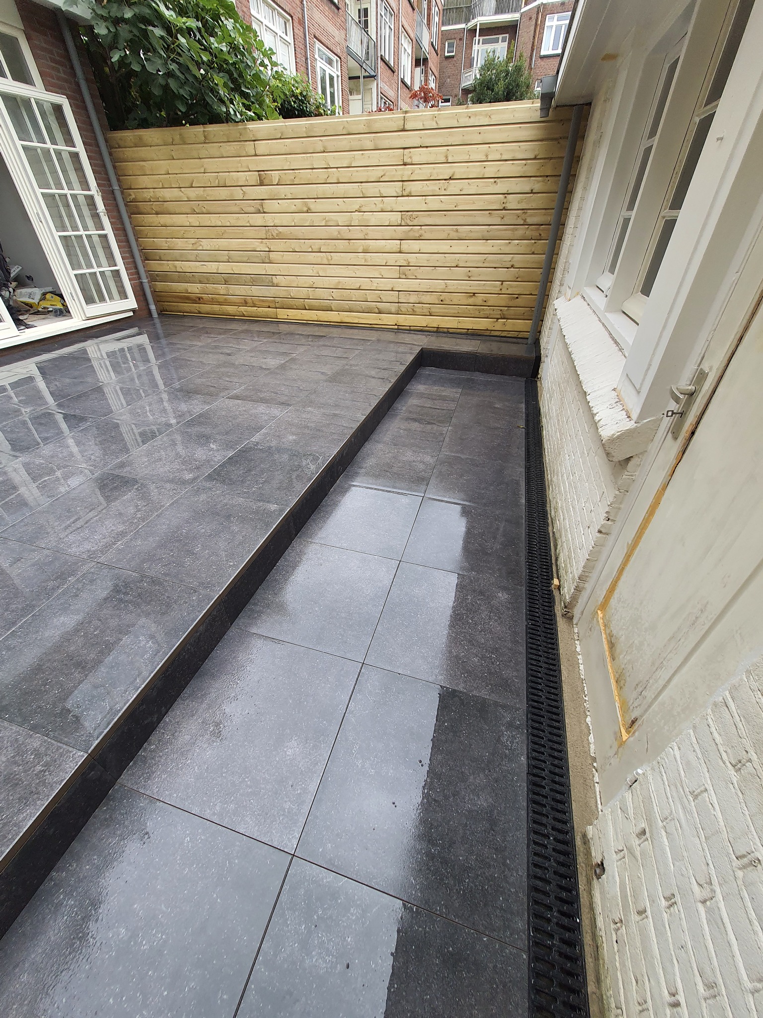 Wyłożony szarymi płytkami taras z drewnianym ogrodzeniem, widoczne okno i odpływ liniowy przy ścianie budynku.