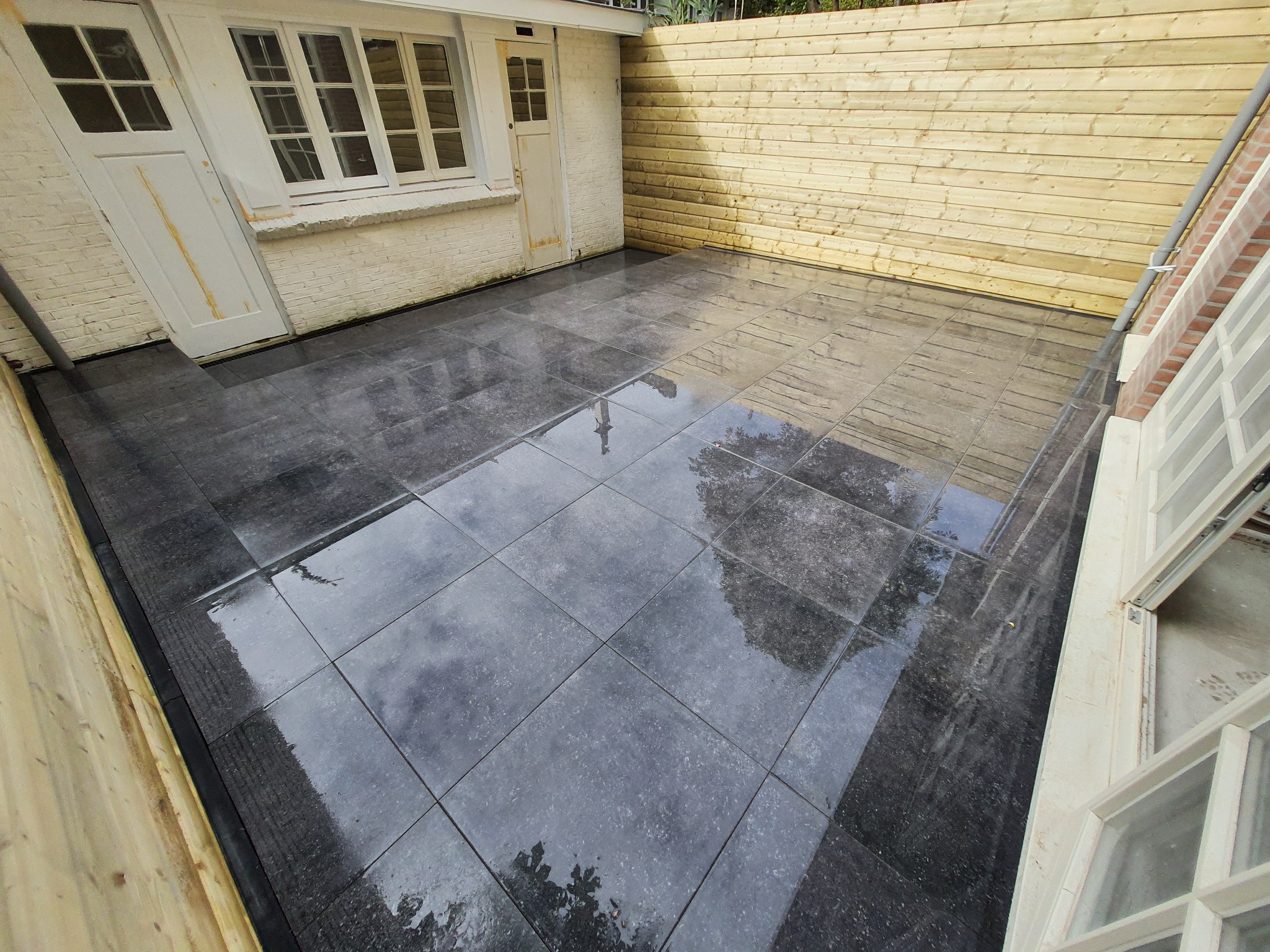 Ułożona, ciemnoszara, kwadratowa podłoga z płytek w małym patio z drewnianym ogrodzeniem i białymi drzwiami z oknem.