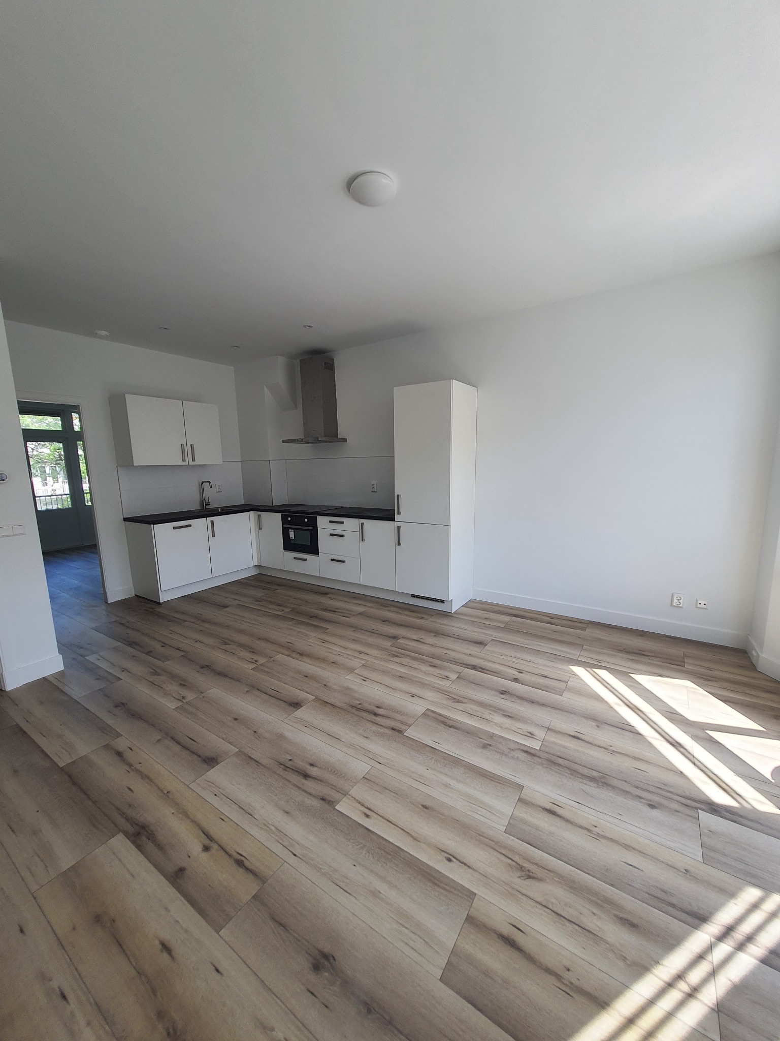 Jasne, przestronne wnętrze z drewnopodobną podłogą, białą zabudową kuchenną z czarnym blatem i stalowym okapem, widok na drzwi balkonowe.