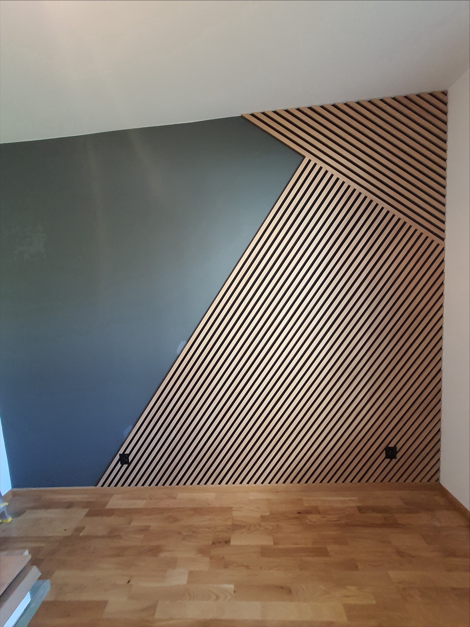 Ściana z drewnianymi lamelami ułożonymi ukośnie, częściowo pomalowana na szaro, z widocznymi gniazdkami elektrycznymi i drewnianą podłogą