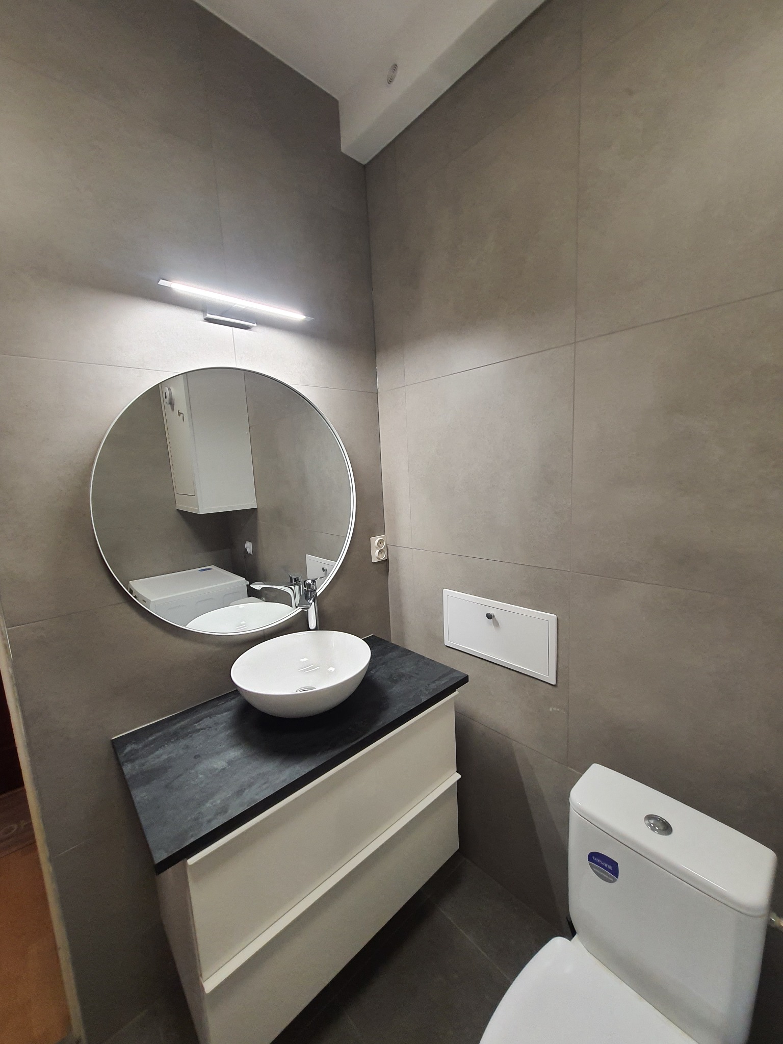 Nowoczesna łazienka z umywalką nablatową na szafce z czarnym blatem, okrągłym lustrem podświetlanym listwą LED i szarą płytką wielkoformatową na ścianach.