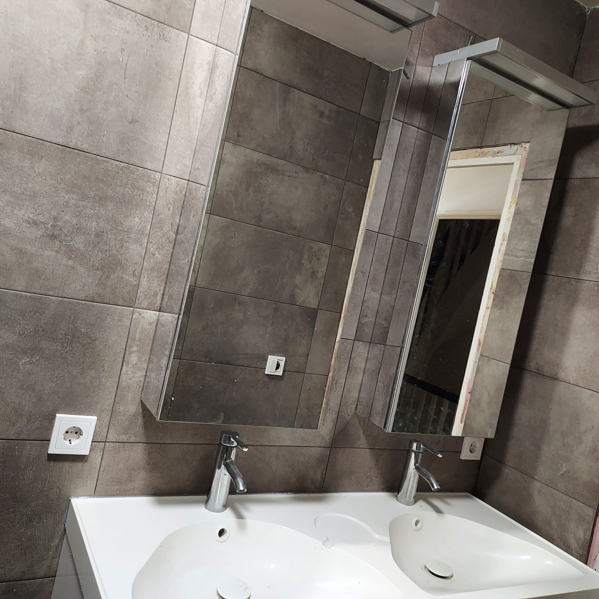 Umywalka z dwoma kranami i lustrami w łazience wyłożonej szarymi, prostokątnymi płytkami; widoczne gniazdko elektryczne na ścianie i fragment instalacji oświetleniowej nad lustrami.