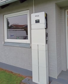 Biały inwerter solarny marki Solax zamontowany na elewacji budynku, z widocznymi przewodami i skrzynką zabezpieczeń, obok okna z odbijającym się w nim białym samochodem dostawczym.