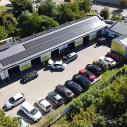 Warsztat samochodowy z panelami słonecznymi na dachu, widok z lotu ptaka, zaparkowane samochody przed budynkiem, jeden z samochodów z otwartą maską.