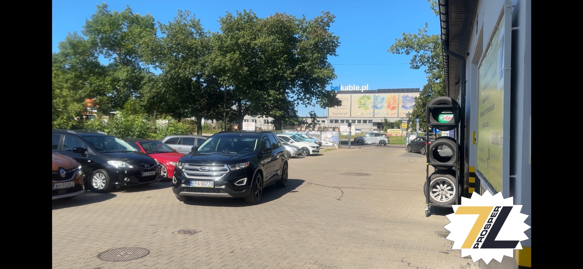 Czarny Ford Edge na parkingu warsztatu samochodowego, widoczne zaparkowane inne auta, w tle budynek z reklamą, obok regał z oponami.