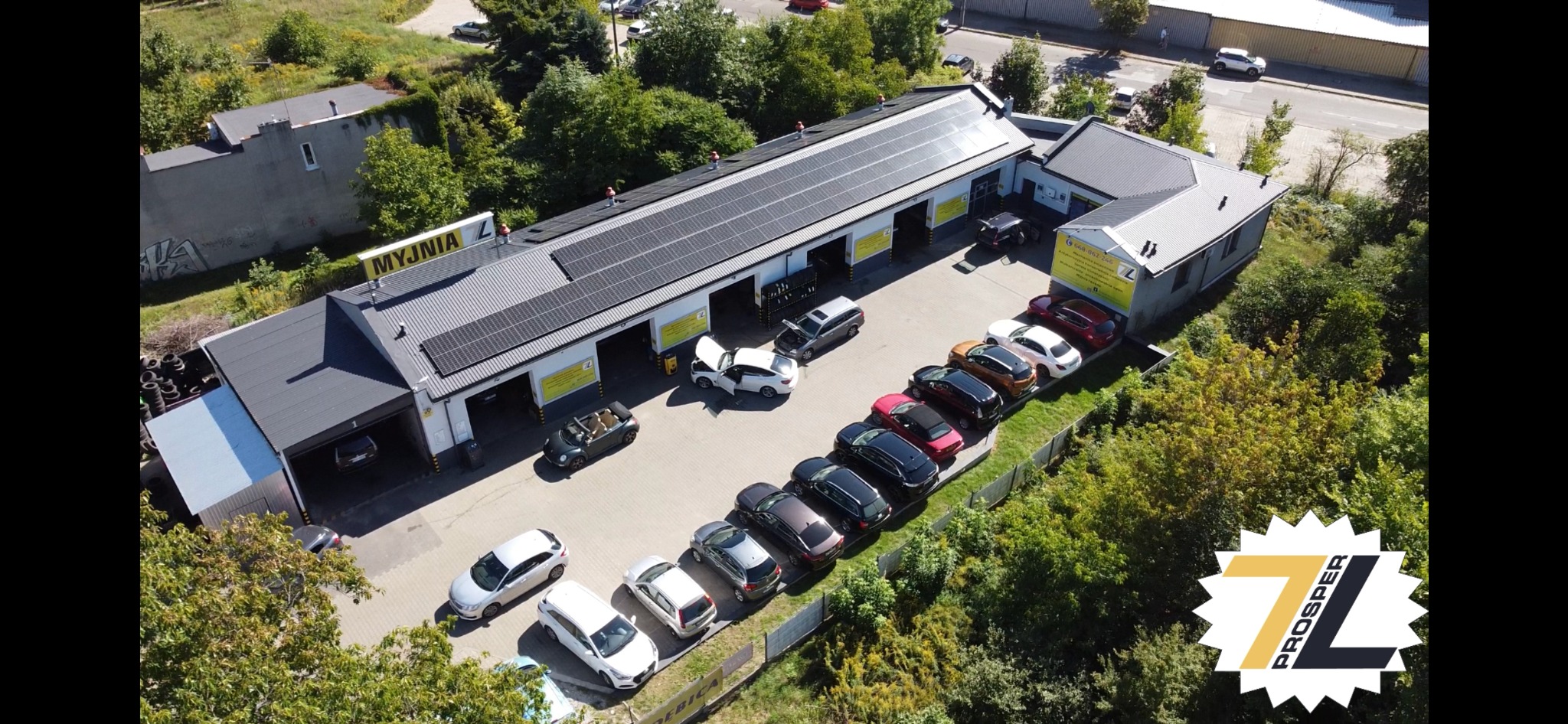 Warsztat samochodowy z panelami słonecznymi na dachu, widok z lotu ptaka, zaparkowane samochody przed budynkiem, jeden z samochodów z otwartą maską.