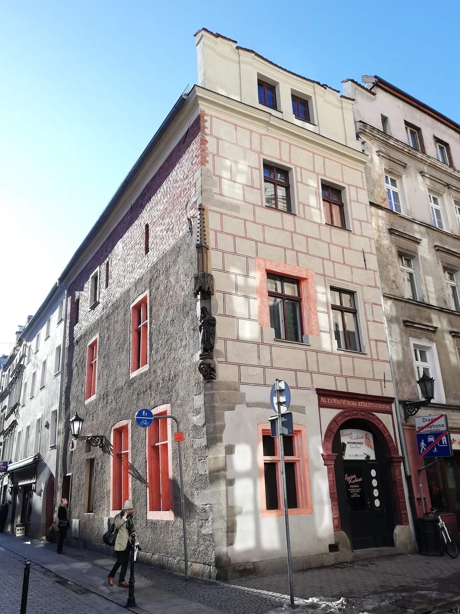 Odnowiona kamienica narożna z dekoracyjnymi elementami w kolorze czerwonym, widoczna z poziomu ulicy z przechodniami i znakami drogowymi.