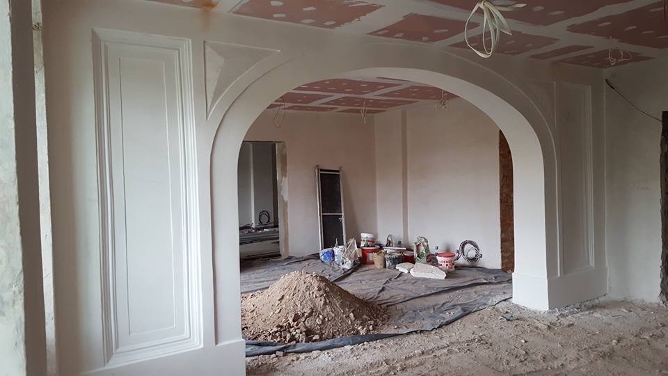 Wnętrze w trakcie prac wykończeniowych: widoczny łuk między pomieszczeniami, ściany po nałożeniu gładzi, przygotowane pod malowanie, oraz góra piasku na podłodze zabezpieczonej folią.