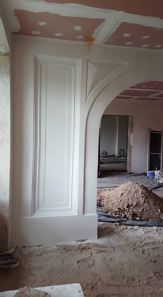 Wnętrze w trakcie prac wykończeniowych: dekoracyjny portal drzwiowy z gładzią, widoczne ślady prac na suficie i podłodze, w tle sterta gruzu i narzędzia.