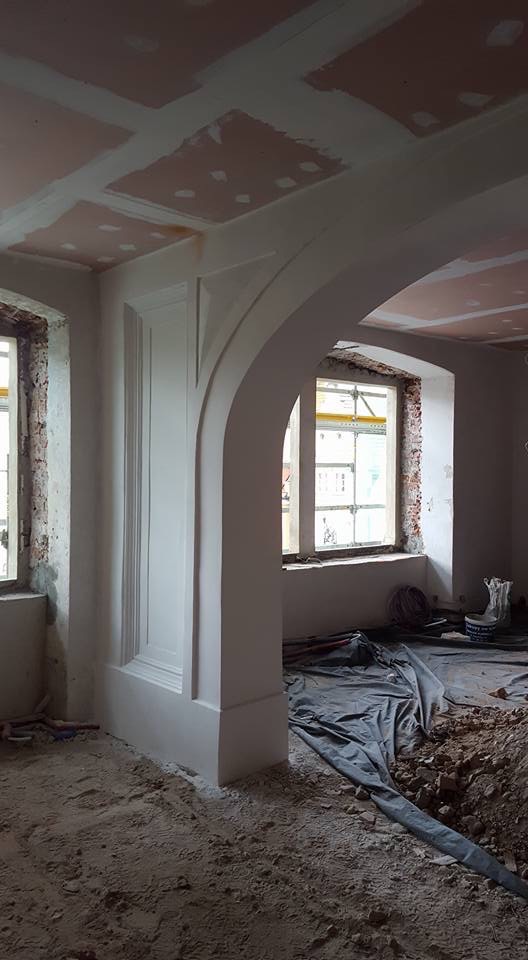 Wnętrze w trakcie remontu: widoczne elementy architektoniczne (łuk, obramowanie), surowy stan ścian i sufitu z widocznymi śladami prac wykończeniowych, podłoga częściowo zakryta folią ochronną.