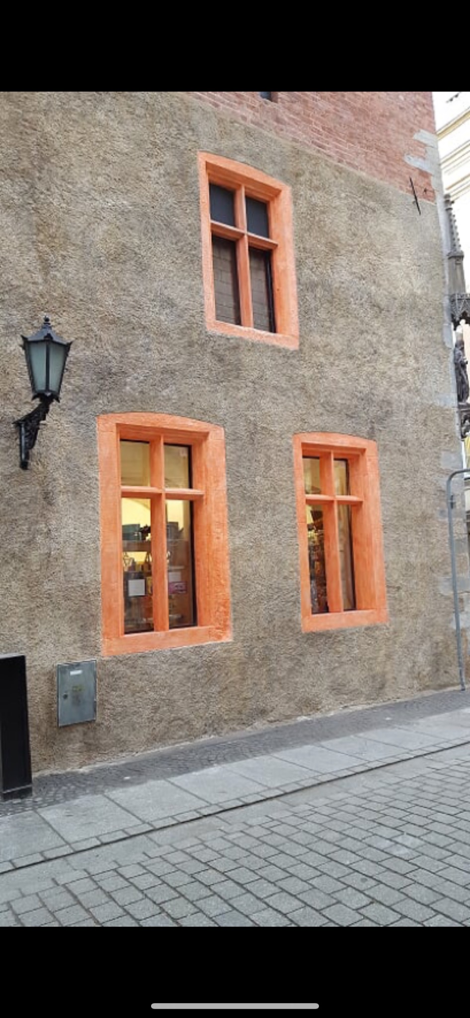 Szara elewacja budynku z trzema oknami w pomarańczowych ramach, widoczna latarnia uliczna i brukowana ulica.