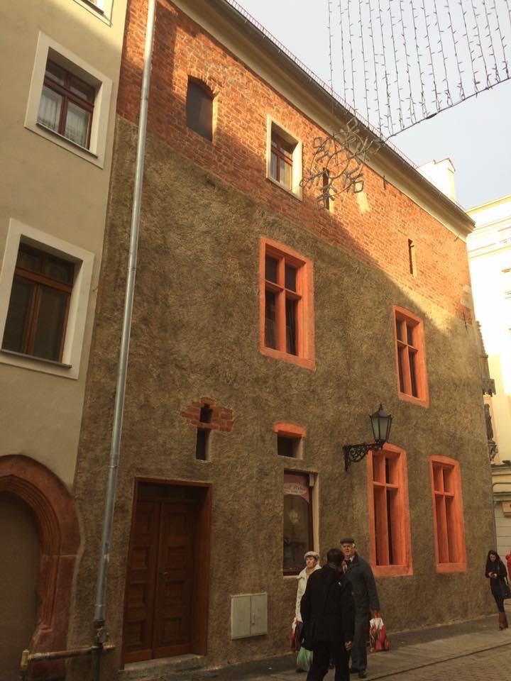 Kamienica z elewacją w odcieniach brązu i cegły, z pomarańczowymi obramowaniami okien i drzwi, widoczne osoby na chodniku.