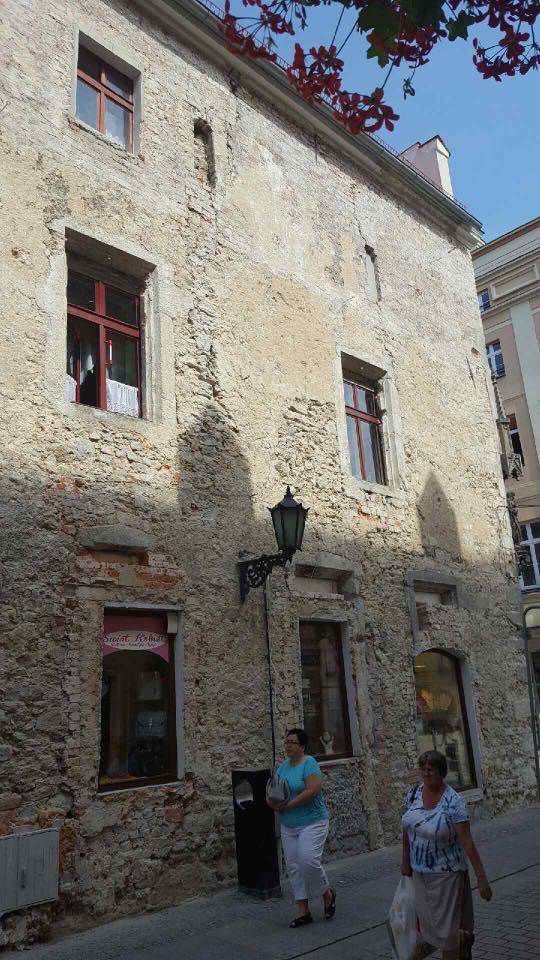 Kamienica z nierówną, zniszczoną elewacją w odcieniach beżu i szarości, z czerwonymi ramami okiennymi, widoczne dwie przechodzące osoby i wisząca latarnia.