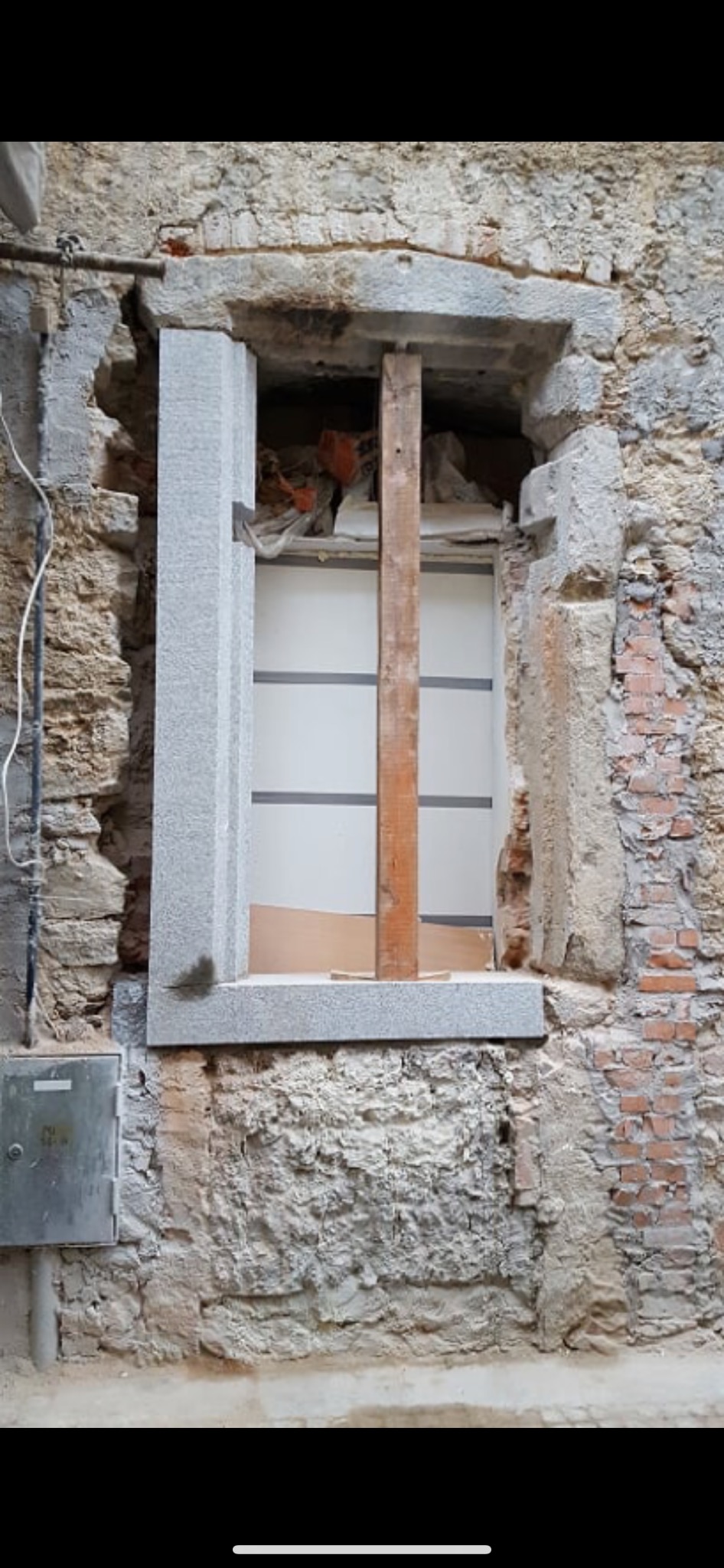 Montaż ościeżnicy w surowej elewacji z widocznym wspornikiem drewnianym i fragmentami starej cegły