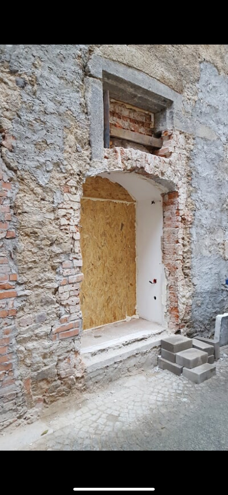 Fragment elewacji budynku w trakcie renowacji, z odsłoniętą cegłą, zamurowanym oknem płytą OSB i przygotowanym wejściem.