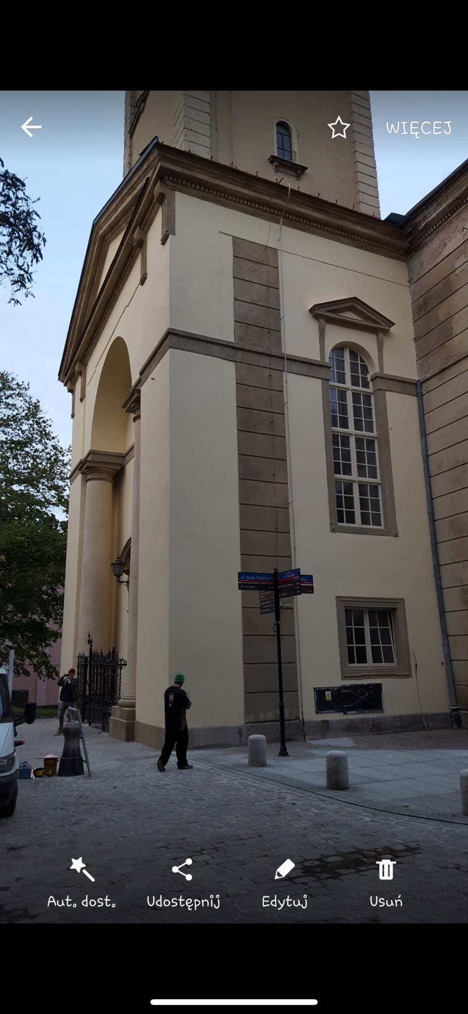 Elewacja budynku sakralnego w trakcie prac renowacyjnych, widoczne rusztowania, pracownicy i elementy architektury.