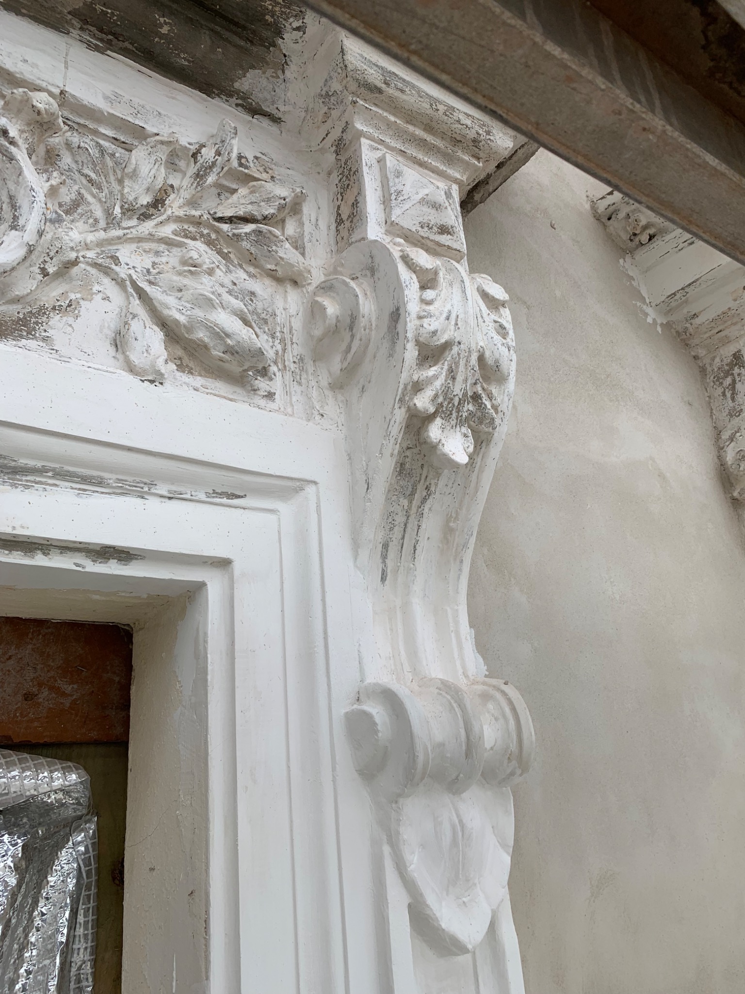 Fragment odrestaurowanej elewacji z dekoracyjnym, białym detalem architektonicznym w stylu klasycznym, widoczne ślady patyny i niedoskonałości, surowy tynk w tle.
