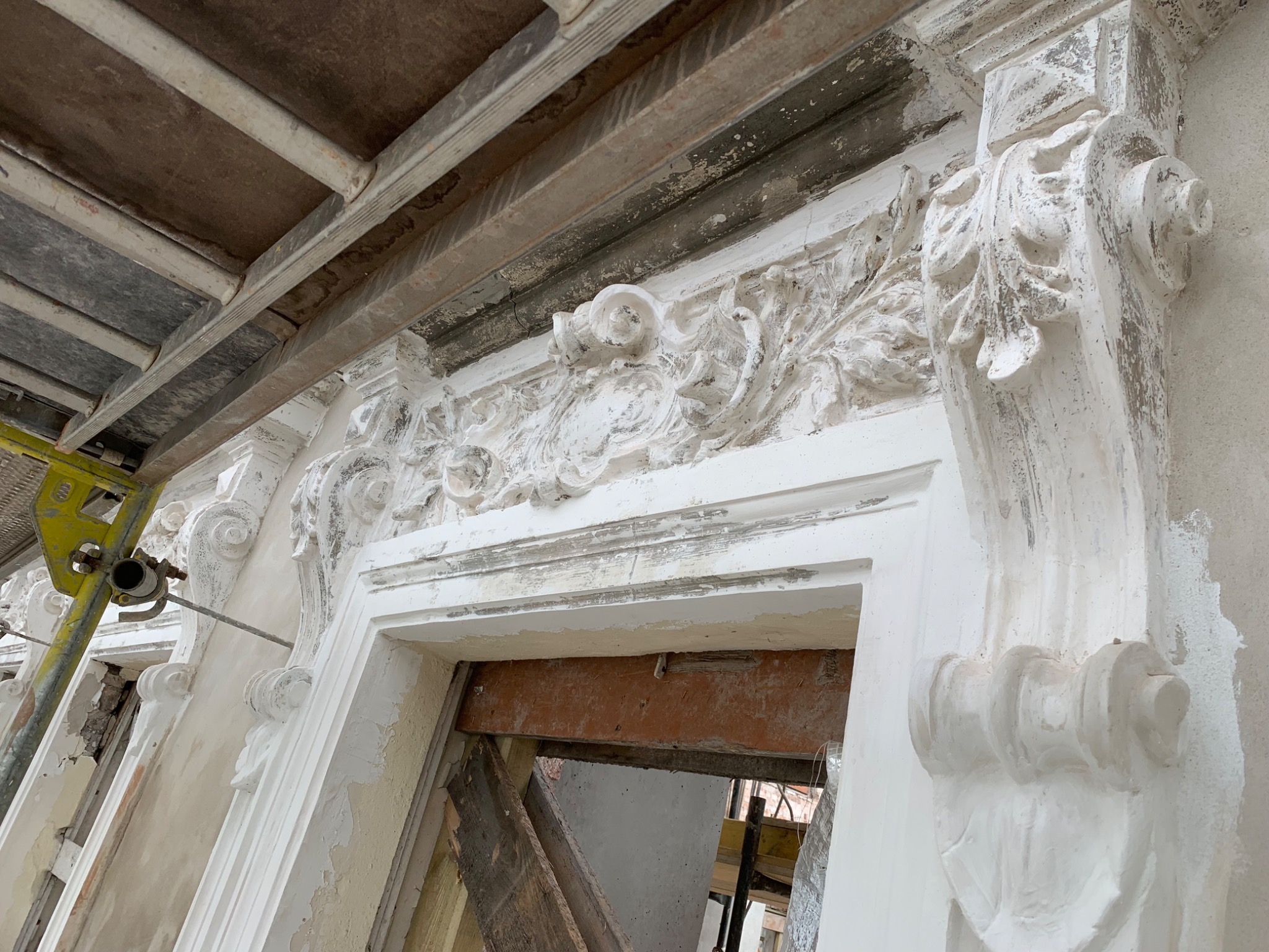 Fragment elewacji budynku z dekoracyjnymi elementami architektonicznymi, w trakcie prac renowacyjnych, widoczne rusztowania i świeżo nałożona biała warstwa, prawdopodobnie tynku lub gładzi.