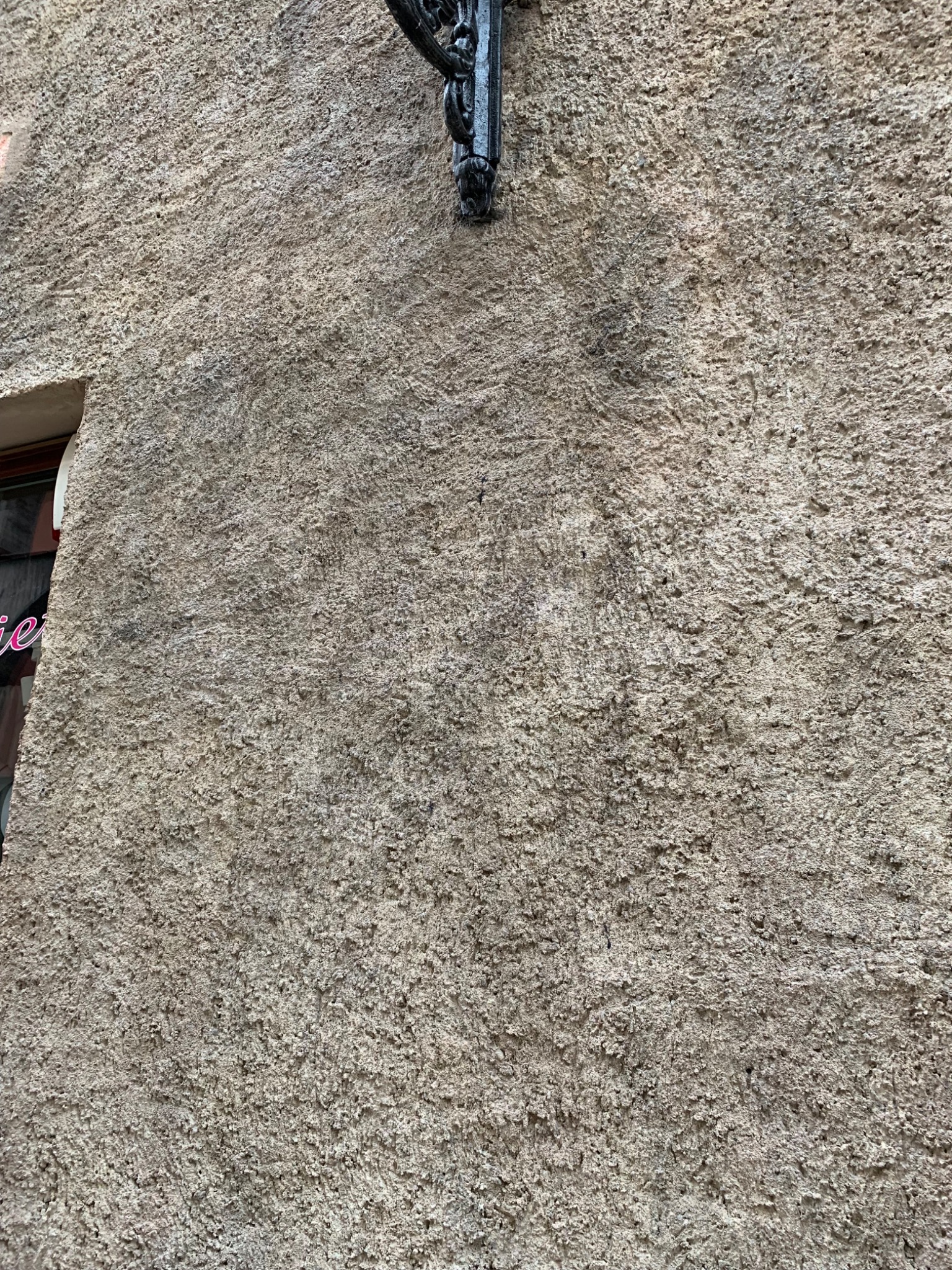 Szorstka, beżowa elewacja budynku z widocznym fragmentem okna i ozdobnym, metalowym wspornikiem lampy.