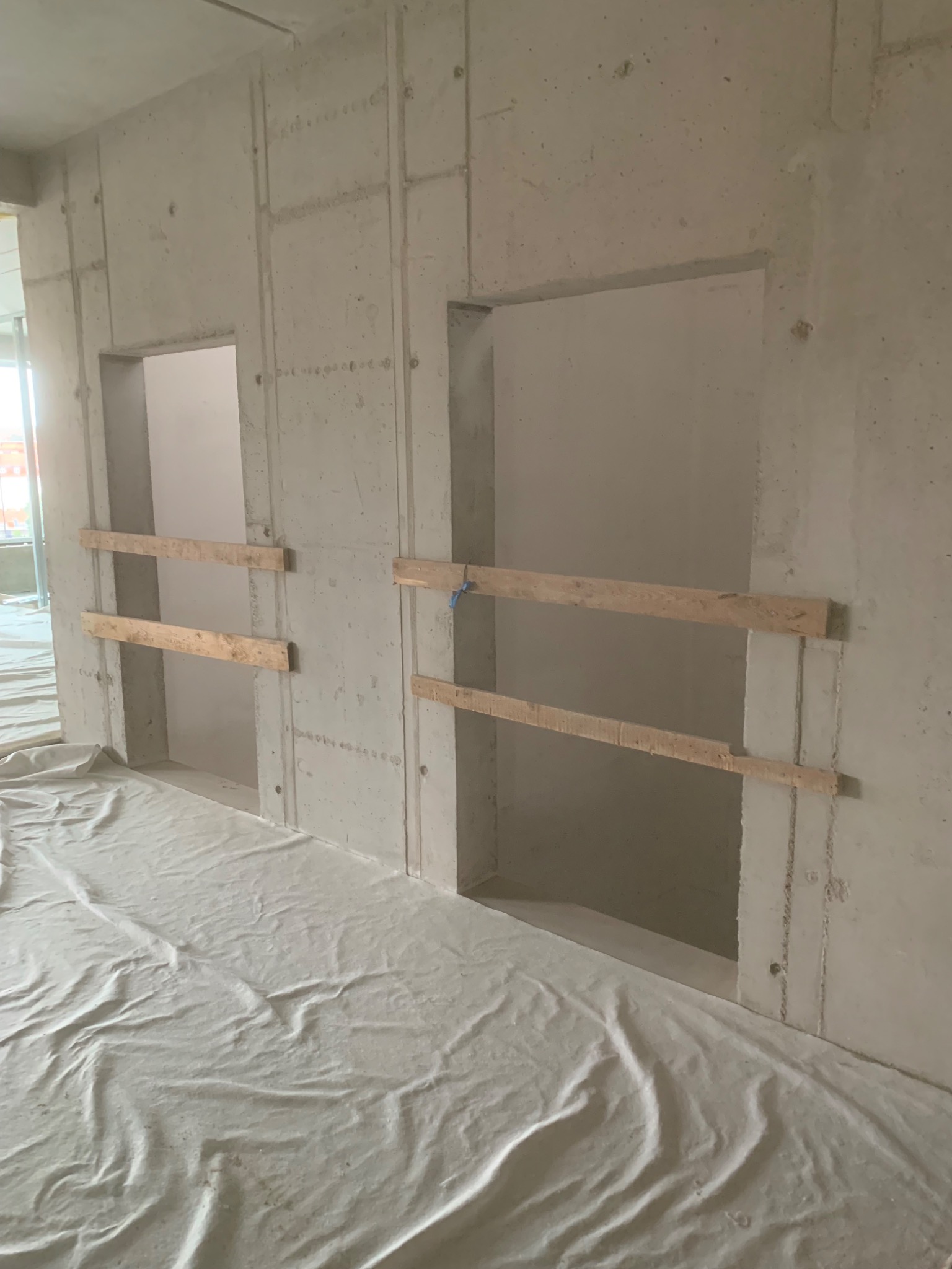 Surowa ściana z betonu architektonicznego z dwoma otworami okiennymi zabezpieczonymi drewnianymi belkami, podłoga przykryta białą folią ochronną, widoczne ślady po szalunkach.