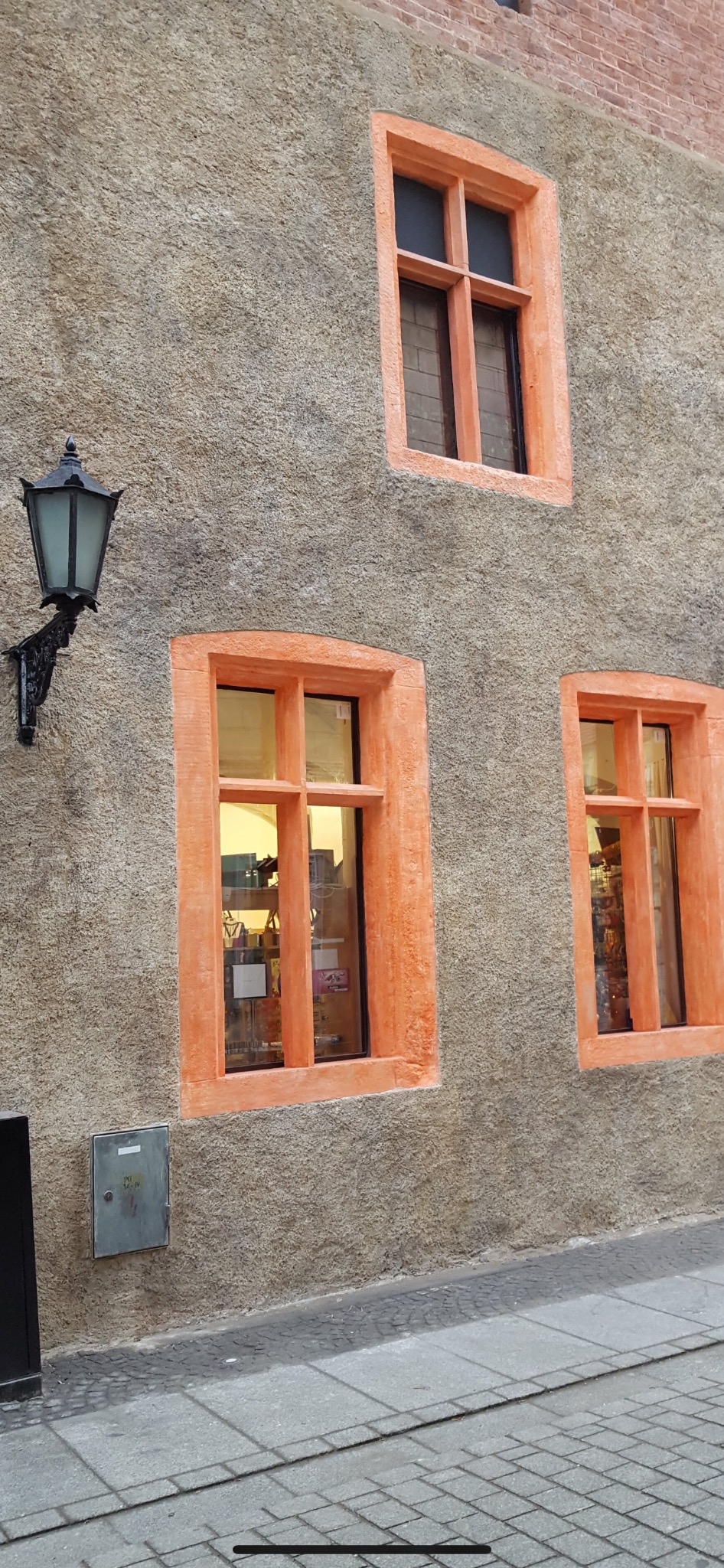 Szara elewacja budynku z trzema oknami o pomarańczowych ramach, widoczna również czarna latarnia i fragment chodnika.