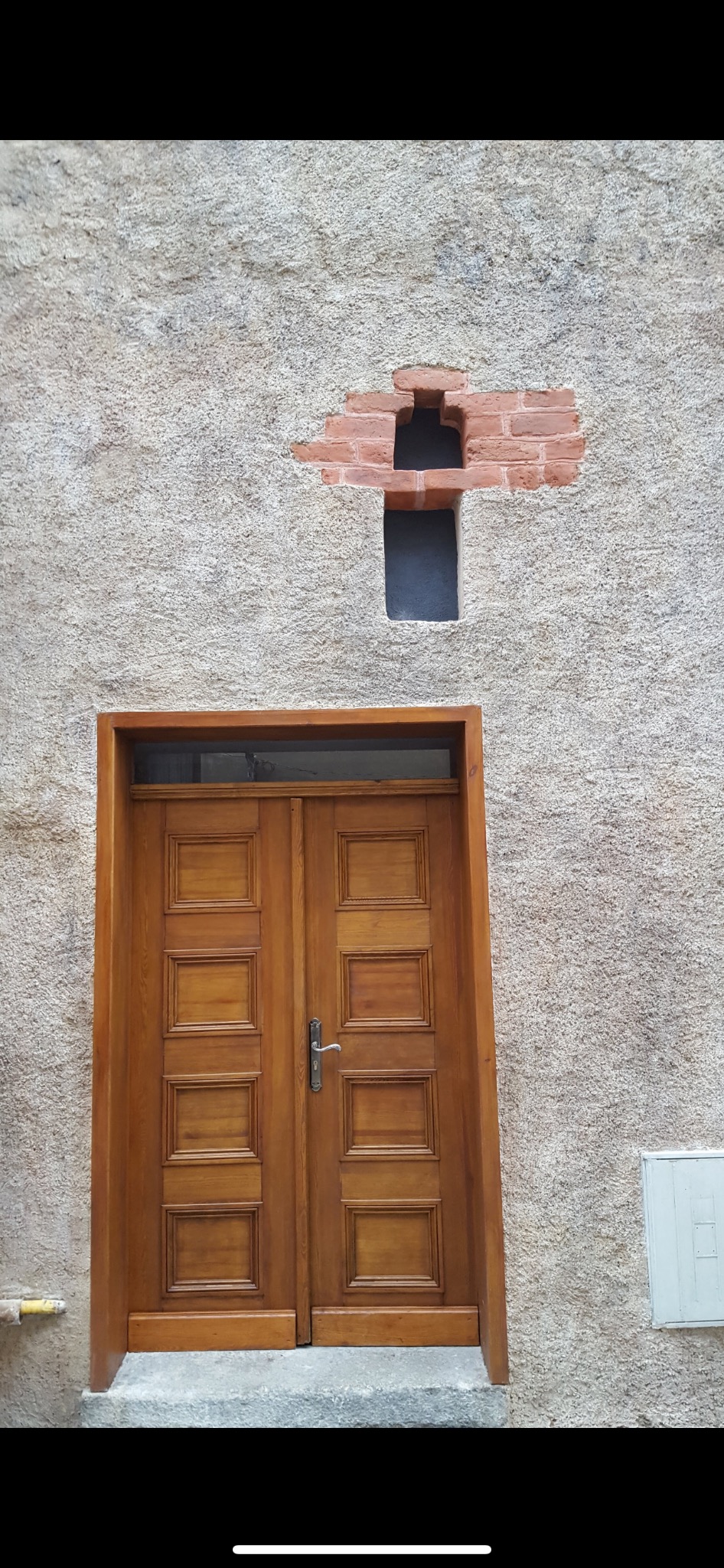 Surowa elewacja z widocznym fragmentem ceglanego otworu wentylacyjnego w kształcie krzyża nad drewnianymi drzwiami wejściowymi.