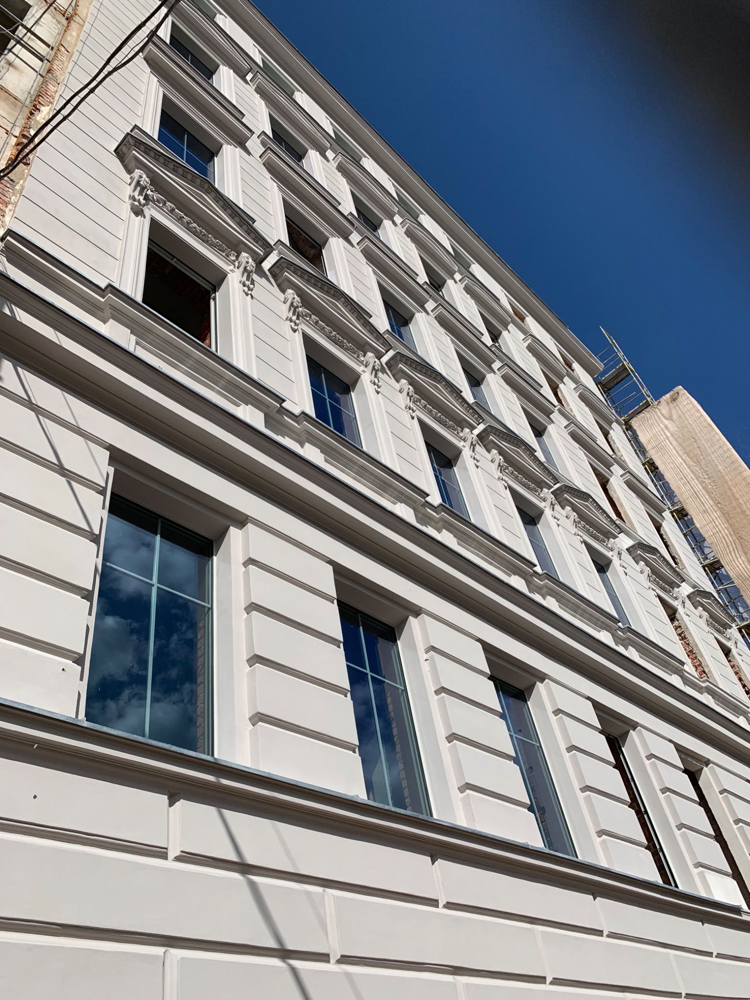 Wysoki budynek z odnowioną, białą elewacją w stylu klasycystycznym, z widocznymi rusztowaniami i fragmentem odsłoniętej cegły na bocznej ścianie, sfotografowany pod kątem w słoneczny dzień.