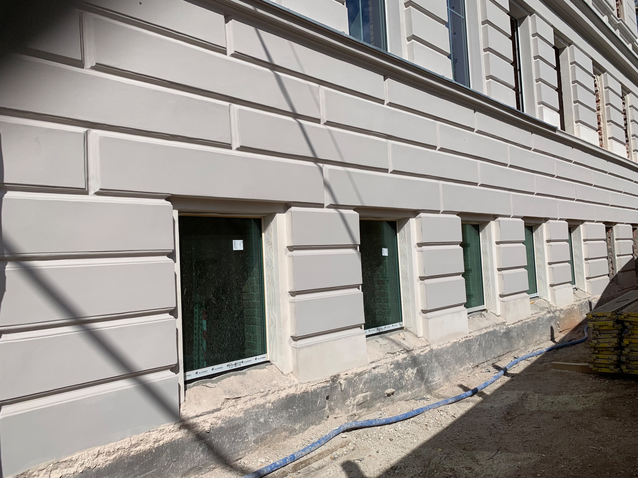 Jasnoszara elewacja budynku z wyraźnymi podziałami na segmenty, widoczne okna z zielonymi szybami, niebieski wąż na ziemi i stos żółtych elementów szalunkowych.