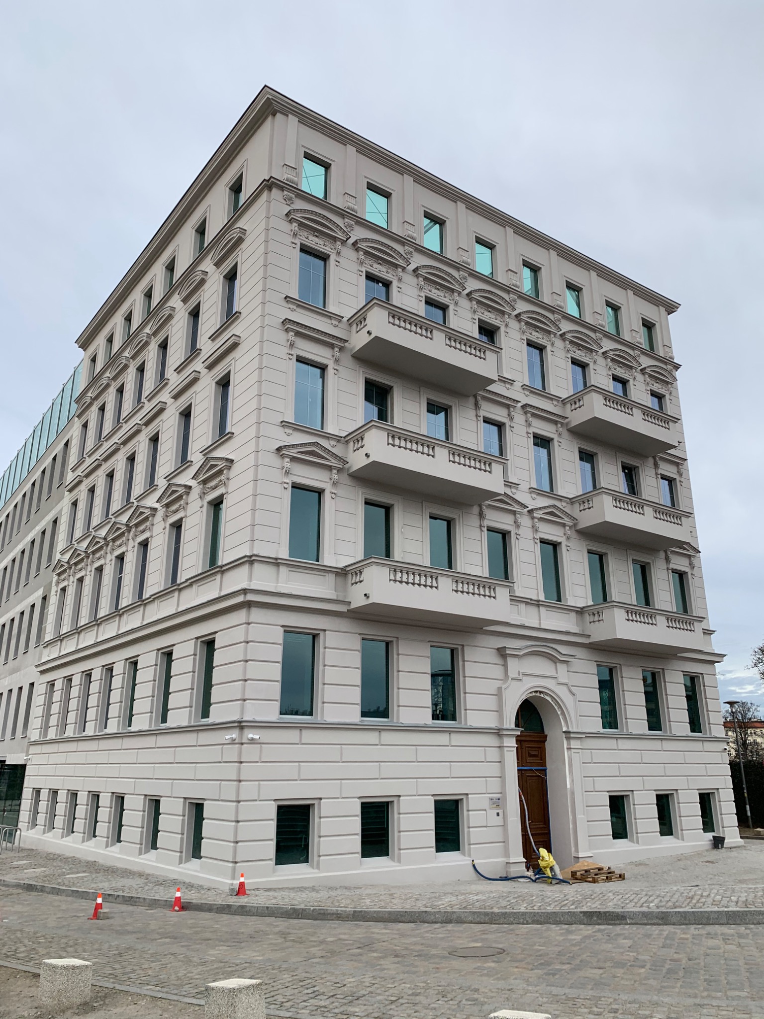 Odnowiona, jasna elewacja budynku z balkonami, widoczne detale architektoniczne i częściowo zakryte wejście z paletami i wężem do czyszczenia.