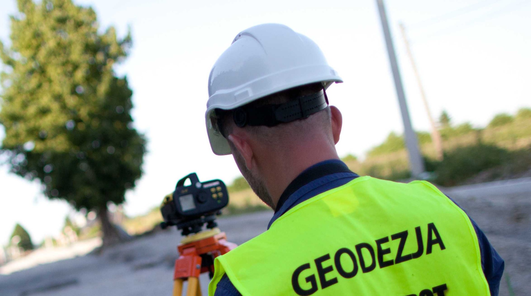 Mężczyzna w kasku i kamizelce odblaskowej z napisem 'GEODEZJA' ustawia teodolit na trójnogu na tle drzew i drogi.