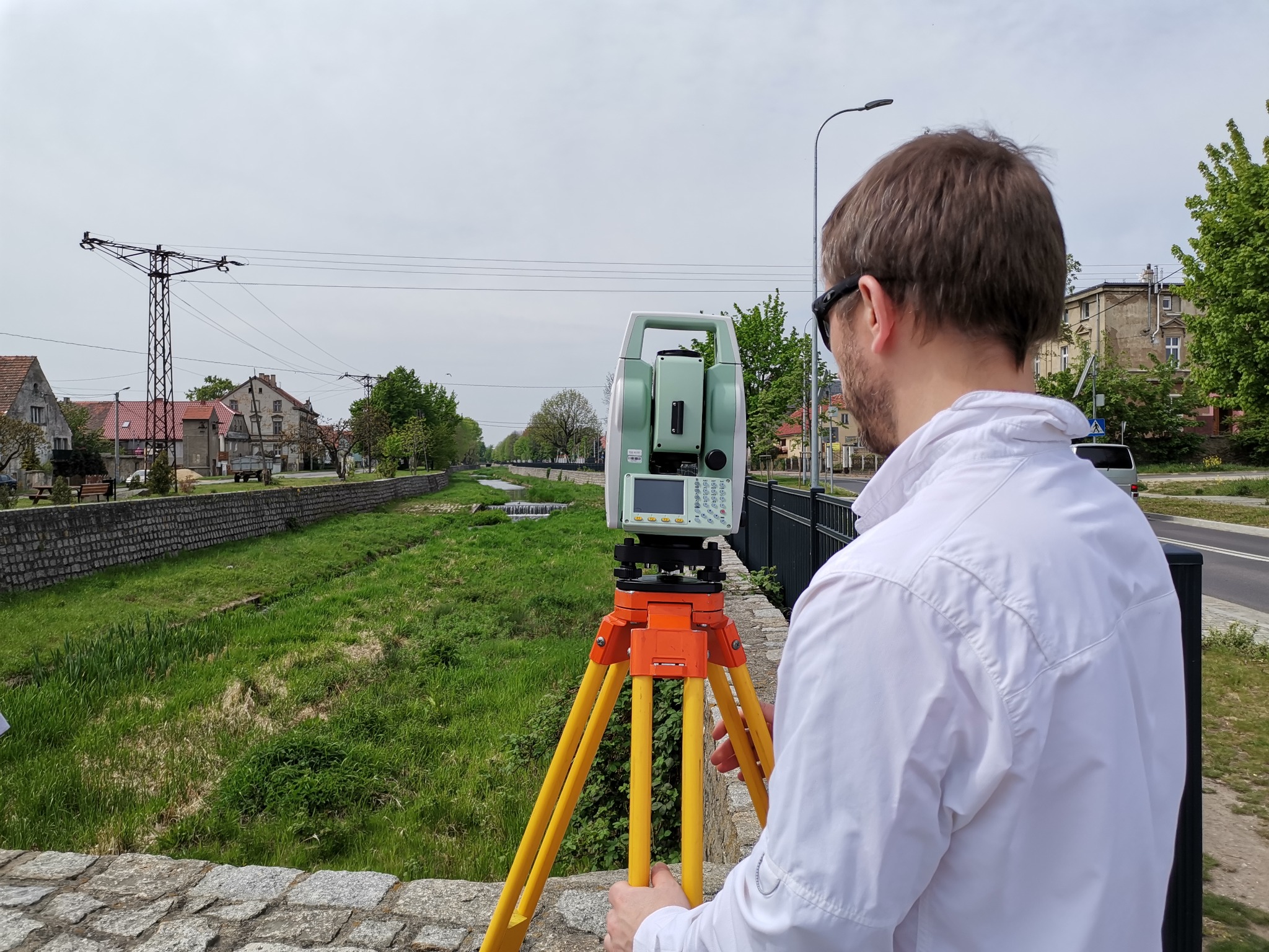 Praca z teodolitem na tle miejskiego krajobrazu z rzeką i zielenią, widok zza pleców osoby w białej koszuli.