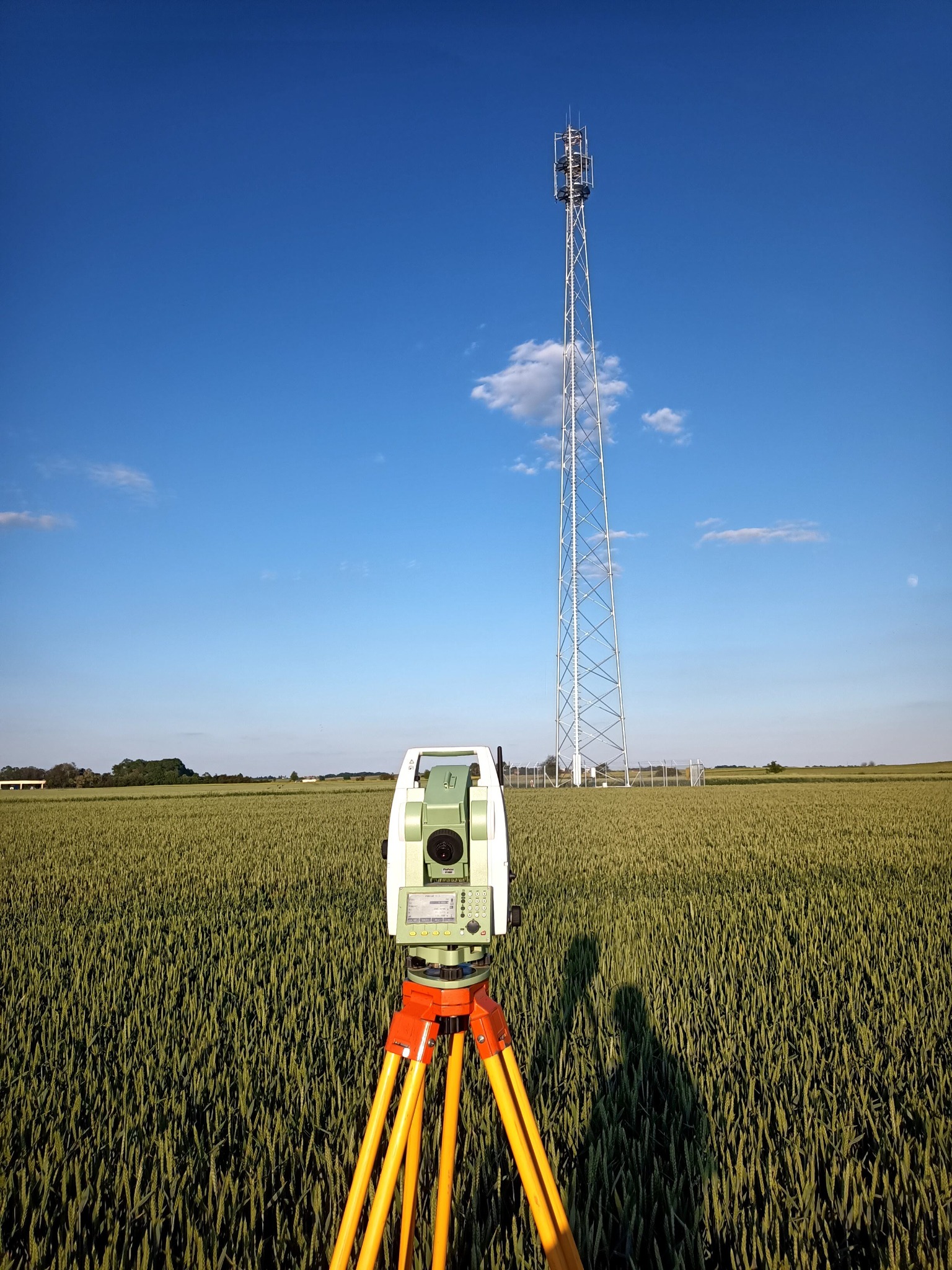 Teodolit na statywie z pomarańczowymi nogami ustawiony na polu zboża, w tle wysoka wieża telekomunikacyjna pod błękitnym niebem z pojedynczymi chmurami.