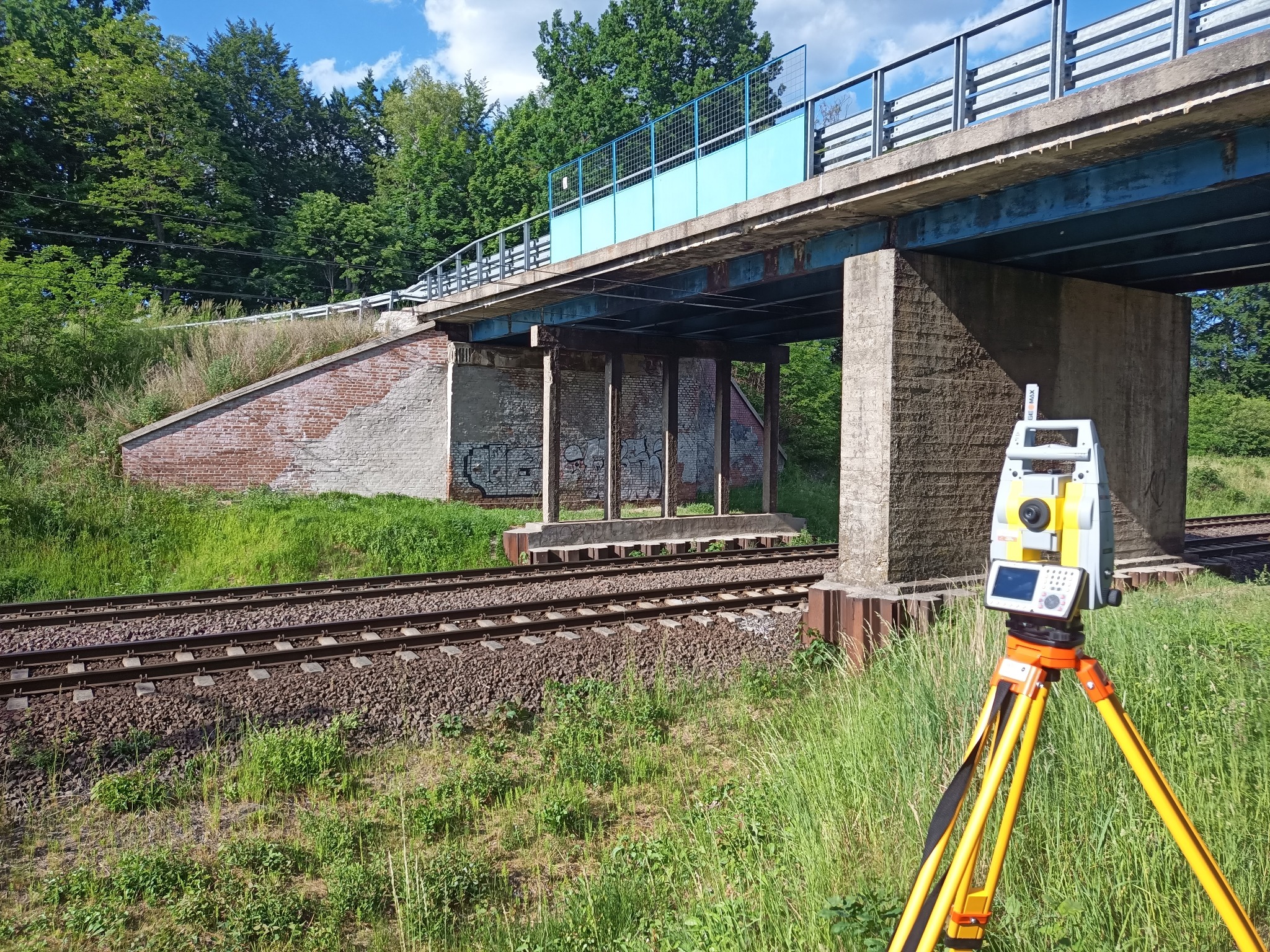 Pomiar geodezyjny torów kolejowych pod wiaduktem z użyciem tachimetru w słoneczny dzień.