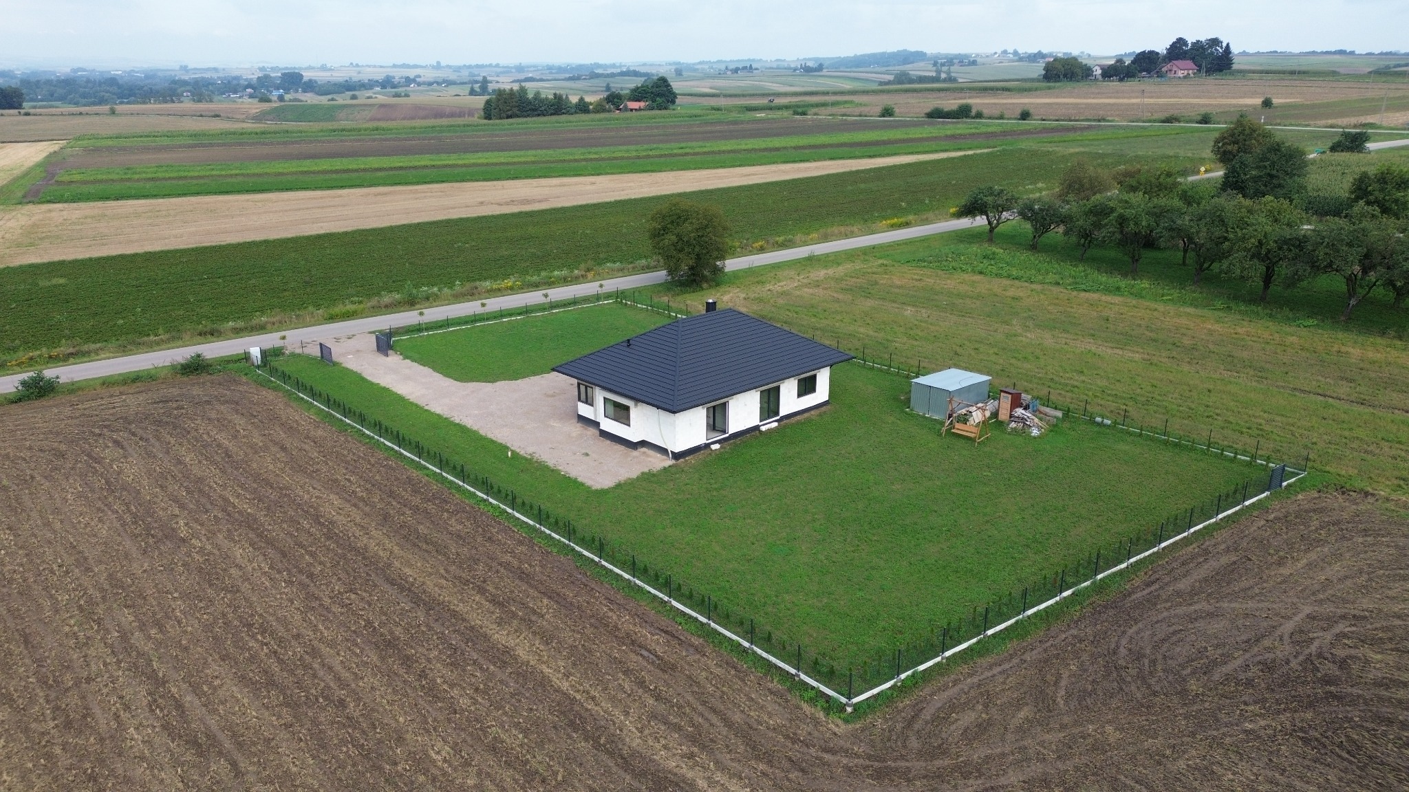 Widok z lotu ptaka na nowoczesny, parterowy dom z ciemnym dachem, otoczony zielonym trawnikiem i ogrodzeniem, z polami uprawnymi w tle.