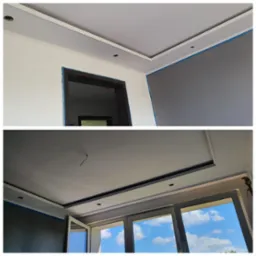 Pomieszczenie w trakcie malowania, widoczne zabezpieczone taśmą malarską obramowania okien i drzwi, ściany w dwóch odcieniach szarości, listwy dekoracyjne pod sufitem.