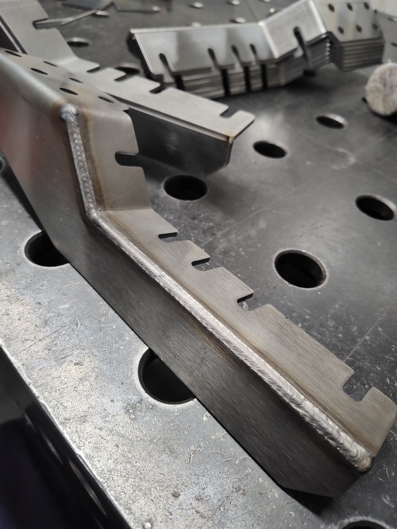 Metalowy profil z precyzyjnym spoiwem spawalniczym na krawędzi, widoczne wycięcia i otwory, ułożony na perforowanym stole spawalniczym.