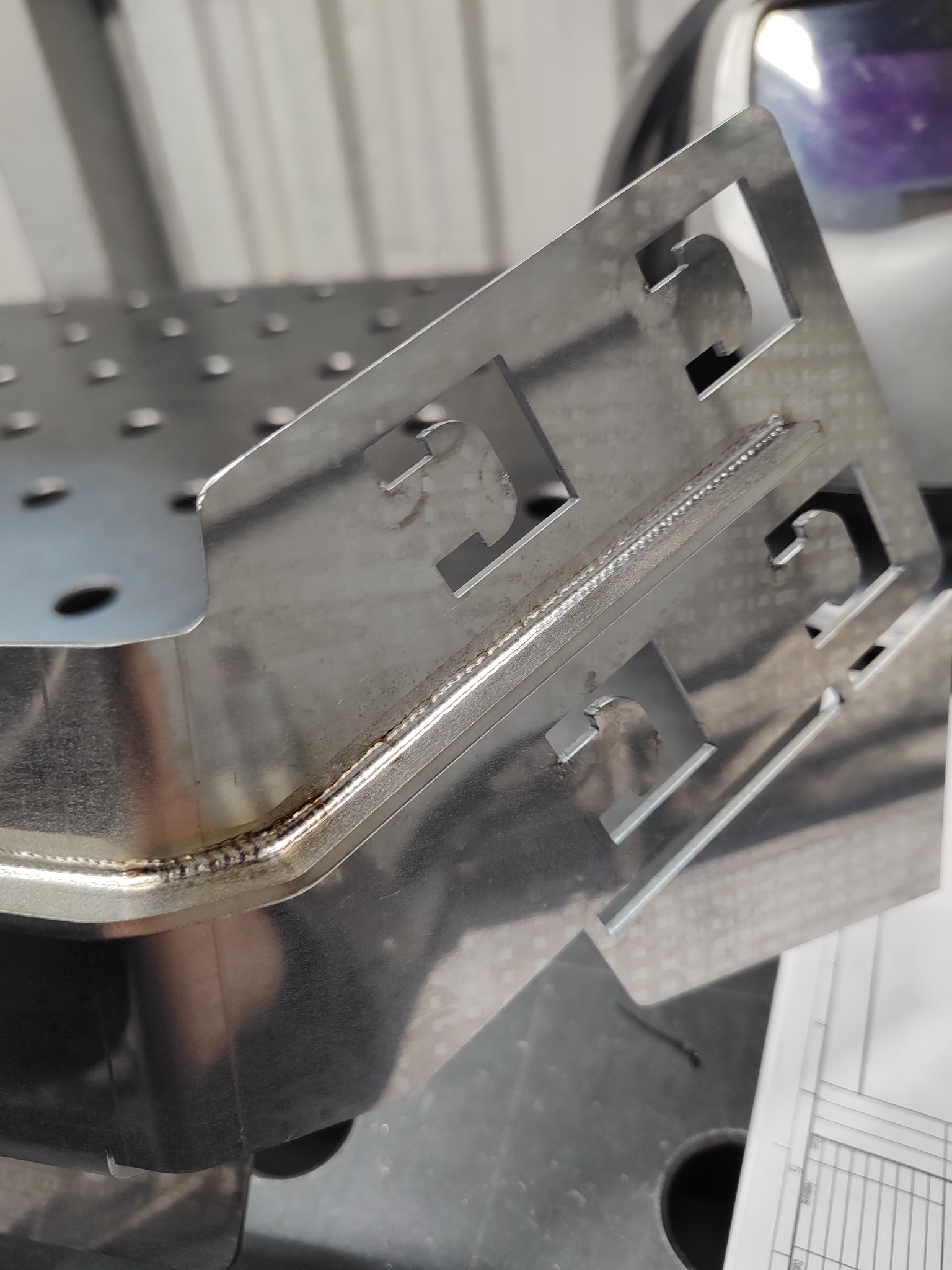 Metalowy element z wyciętymi otworami, spawany na krawędziach, leżący na perforowanym stole spawalniczym; w tle widoczna maska spawalnicza.