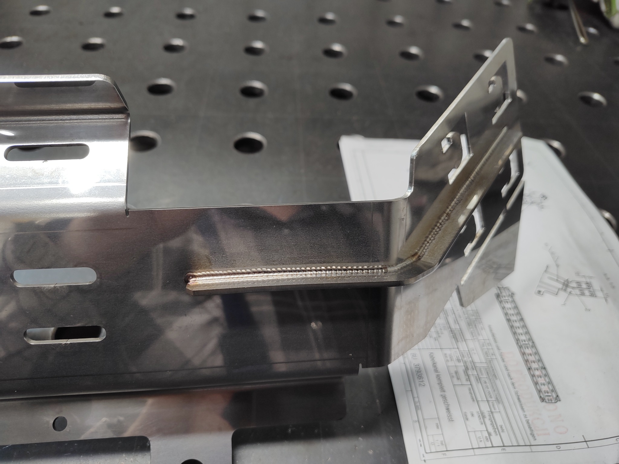 Detal ze stali nierdzewnej, z widocznymi spawami, ułożony na perforowanym stole spawalniczym obok dokumentacji technicznej.
