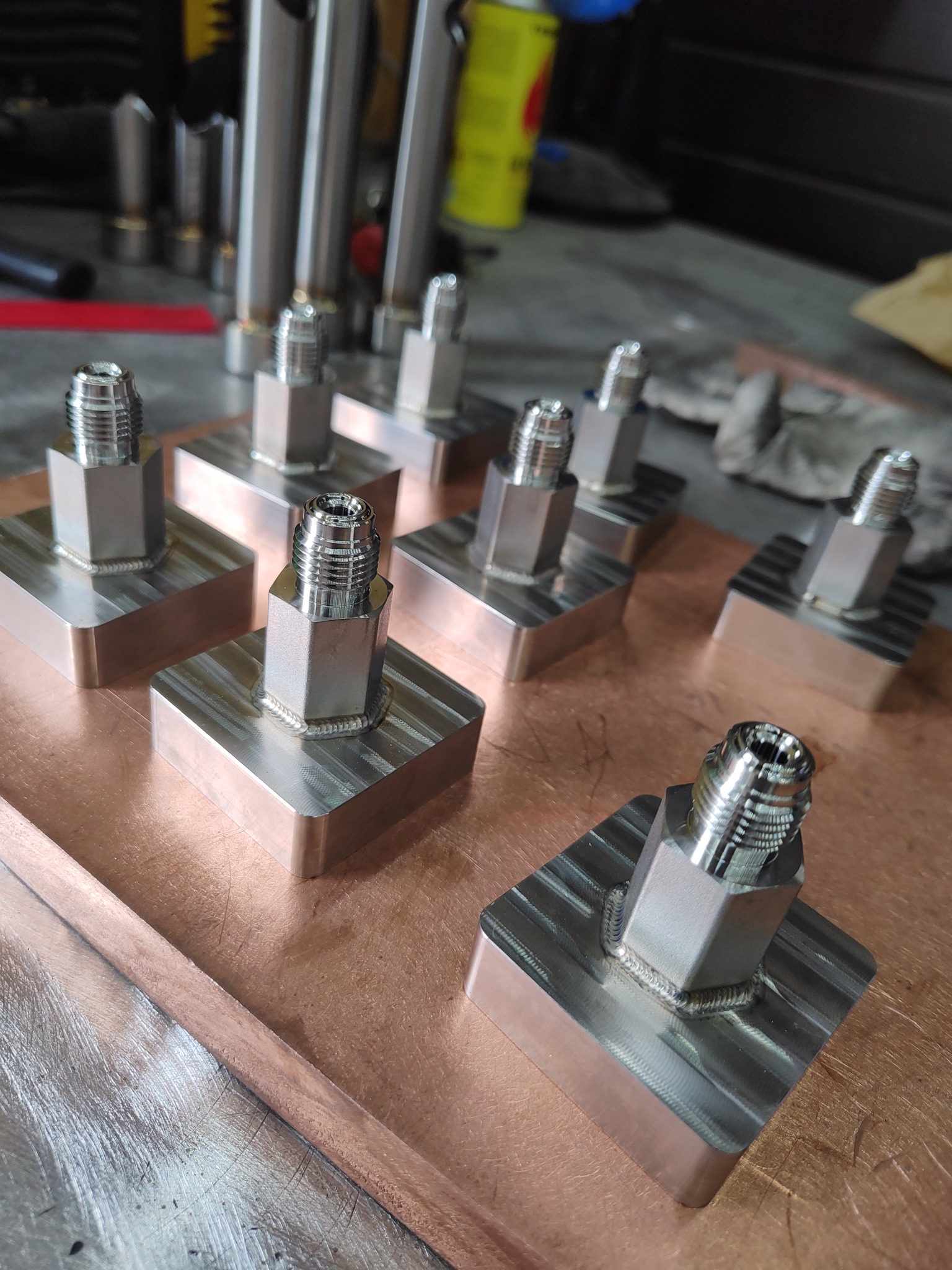 Precyzyjnie wyspawane, chromowane złączki na metalowych podstawach z widocznymi śladami obróbki skrawaniem, ułożone rzędami na miedzianej powierzchni roboczej.