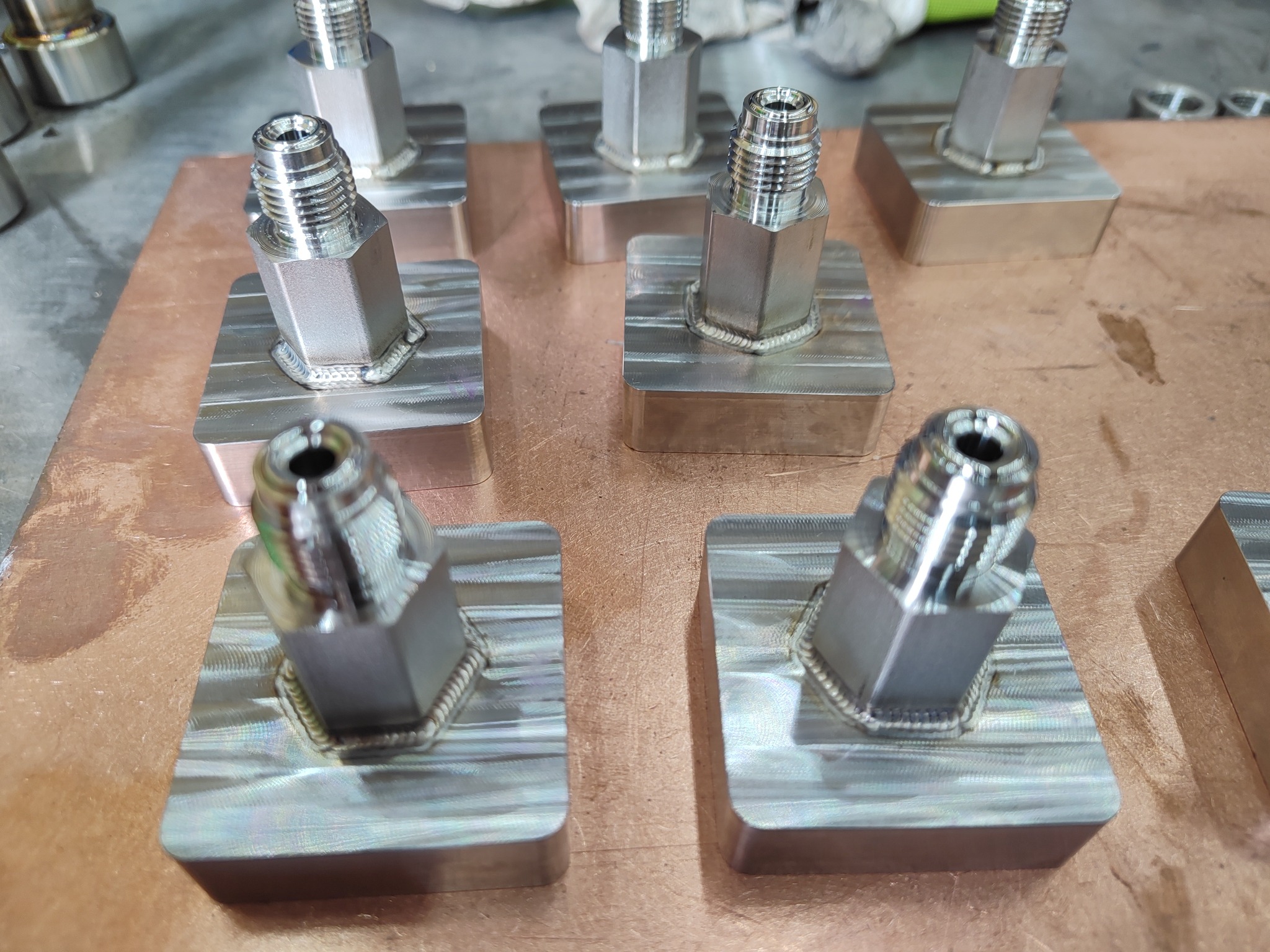 Seria metalowych elementów z gwintem, każdy spawany do kwadratowej podstawy, ułożonych na miedzianej powierzchni roboczej.