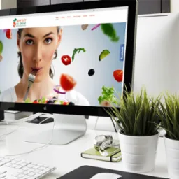 Ekran komputera iMac wyświetla stronę internetową z kobietą jedzącą sałatkę, obok telefon z wersją mobilną strony, na białym biurku z roślinami w doniczkach.