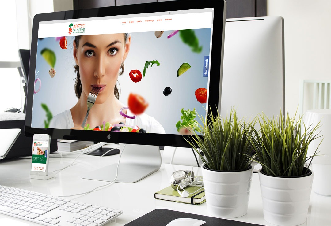 Ekran komputera iMac wyświetla stronę internetową z kobietą jedzącą sałatkę, obok telefon z wersją mobilną strony, na białym biurku z roślinami w doniczkach.