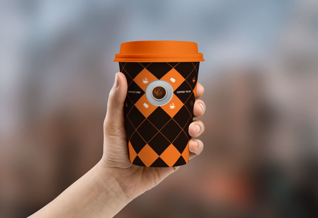 Dłoń trzyma kubek na kawę z pomarańczową pokrywką i wzorem w romby oraz logo kawy na rozmytym tle.