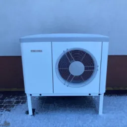 Biała pompa ciepła Stiebel Eltron, przysypana śniegiem, stoi na kostce brukowej również pokrytej śniegiem, na tle jasnej ściany budynku z małym oknem.