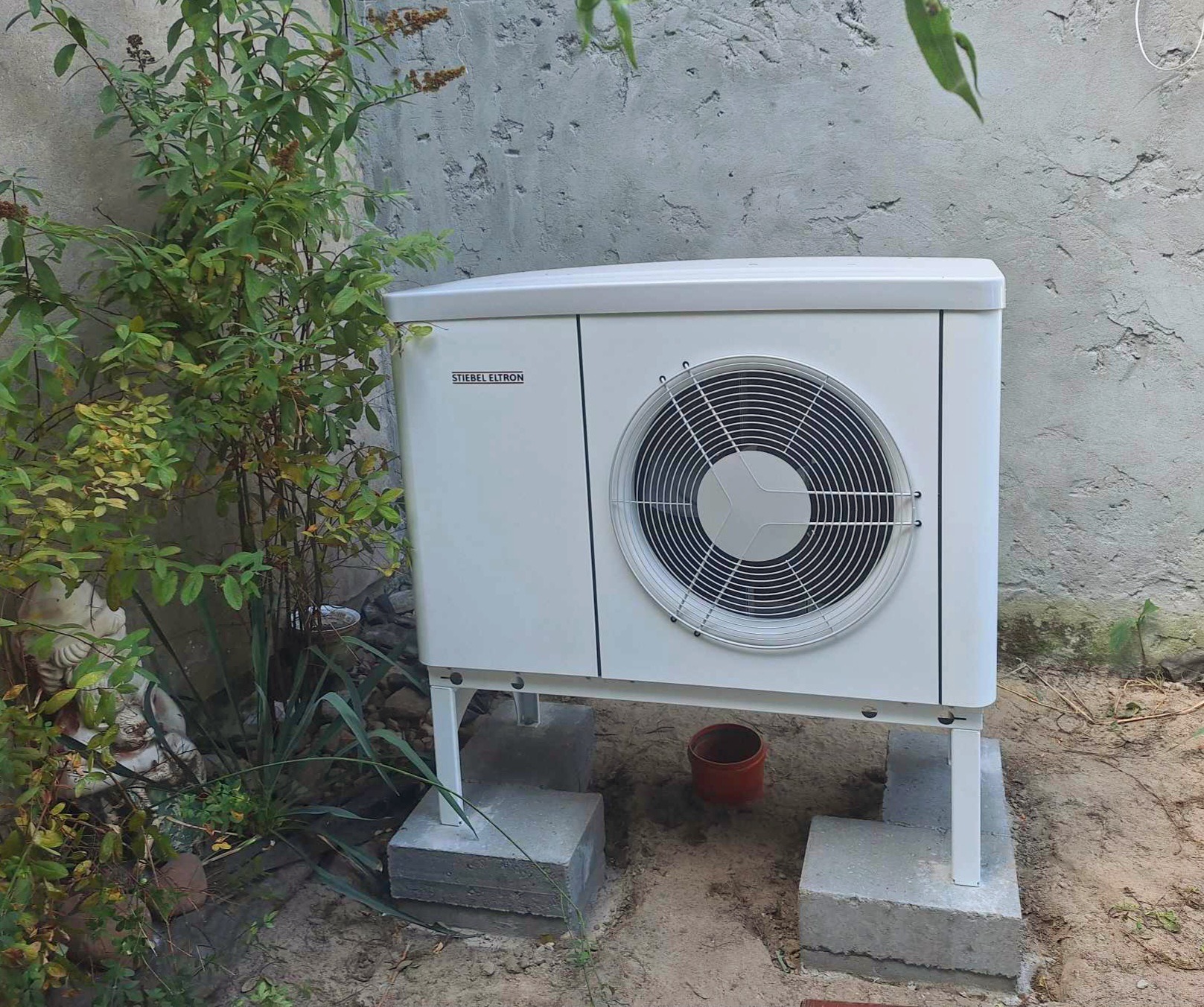 Biała pompa ciepła Stiebel Eltron zamontowana na betonowych podstawach, obok roślinności i rzeźby ogrodowej na tle szarej ściany.