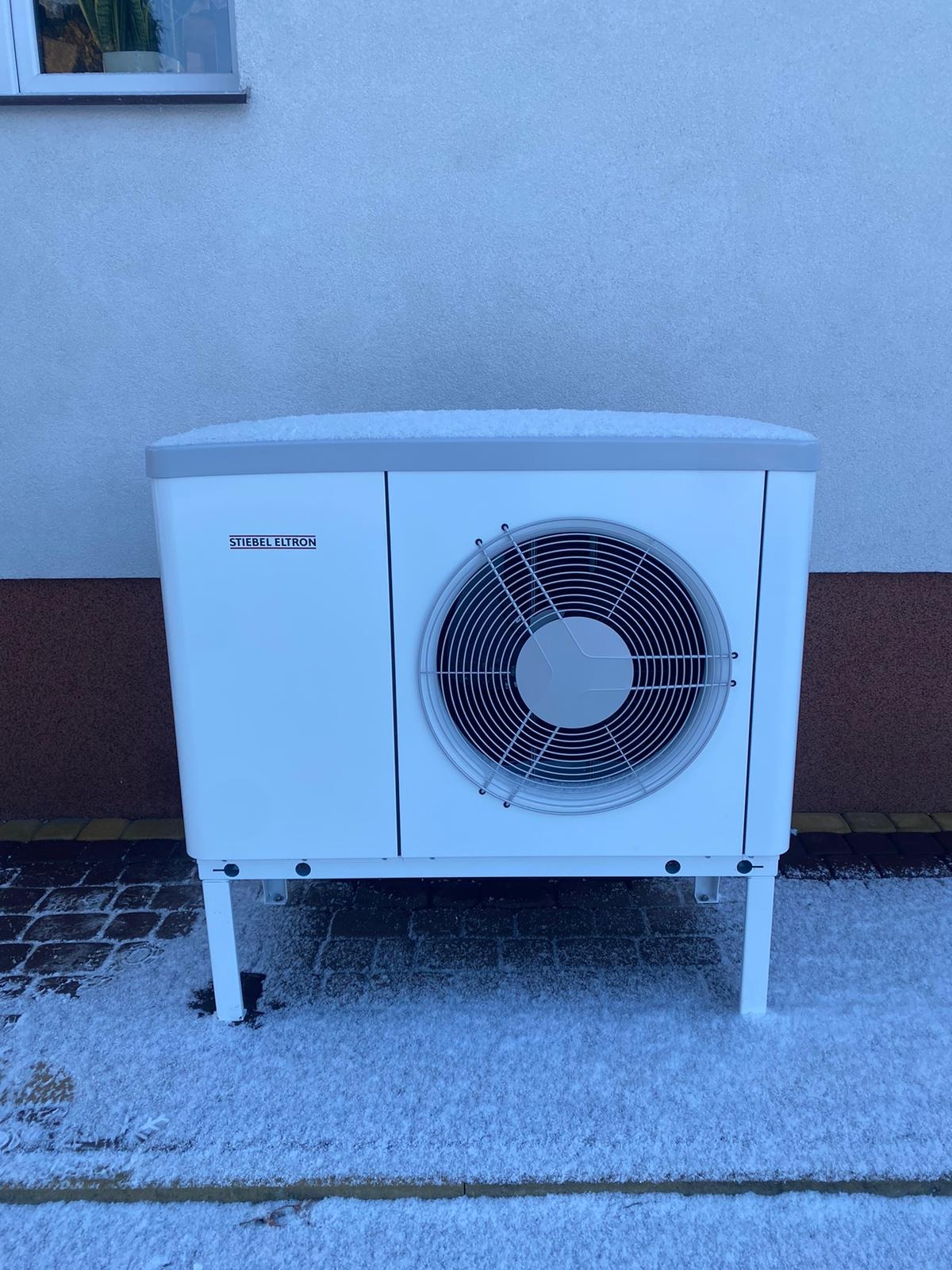 Biała pompa ciepła Stiebel Eltron, przysypana śniegiem, stoi na kostce brukowej również pokrytej śniegiem, na tle jasnej ściany budynku z małym oknem.
