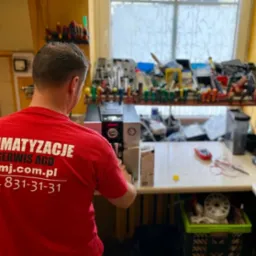 Serwisant AGD w czerwonej koszulce z logo firmy naprawia ekspres do kawy na warsztacie z półką pełną narzędzi.