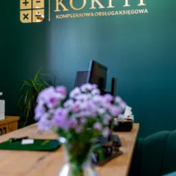 Recepcja biura oferującego kompleksową obsługę finansową, z złotym logo na ciemnozielonej ścianie, bukietem fioletowych kwiatów na drewnianym biurku i widocznymi monitorami w tle.